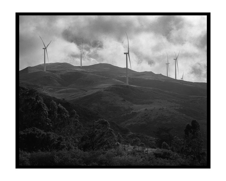 Wind turbines stand on a hilly landscape under a cloudy sky.