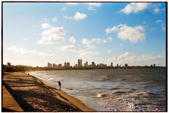 A person stands on a sandy beach next to the ocean with a city skyline in the distance under a cloudy sky.