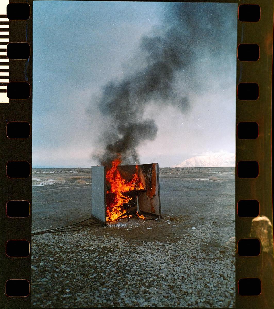 A burning refrigerator emits thick black smoke into the sky in a desolate, rocky landscape with a distant mountain.