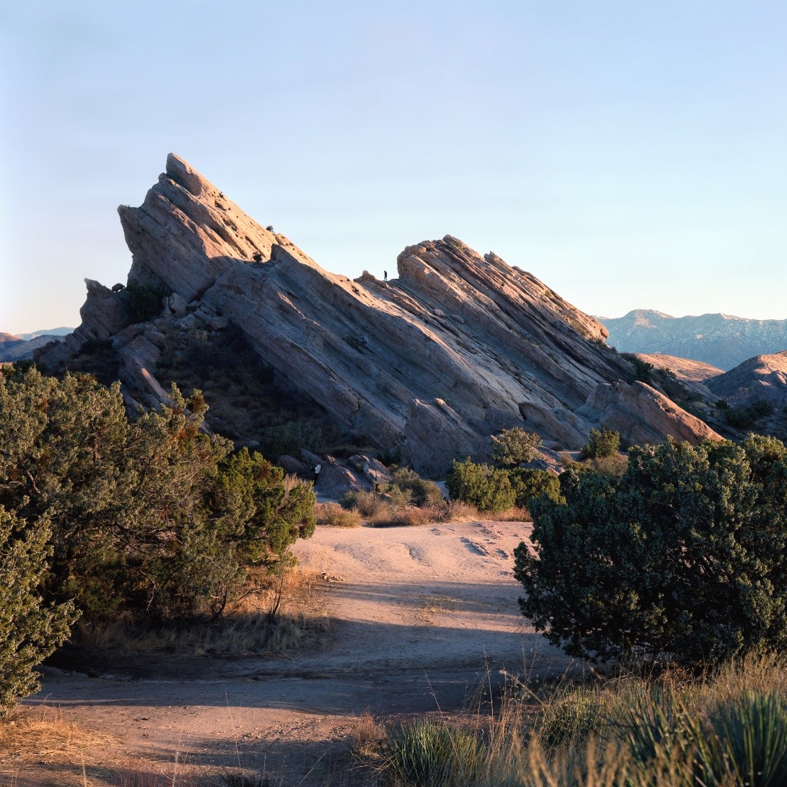 A person stands atop a large, jagged rock formation in a desert landscape with sparse vegetation.