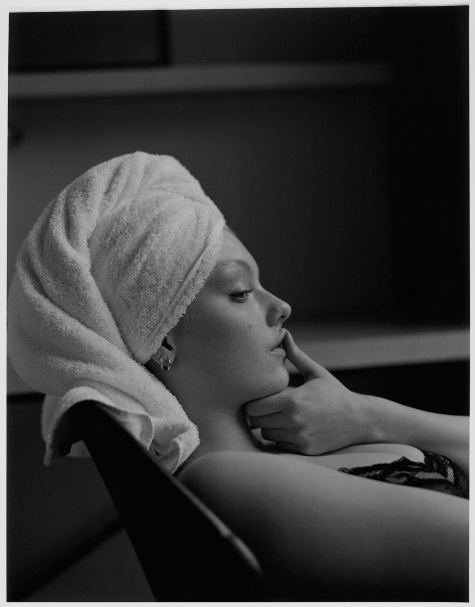 A woman with a towel wrapped around her head rests her chin on her hand and touches her lip.