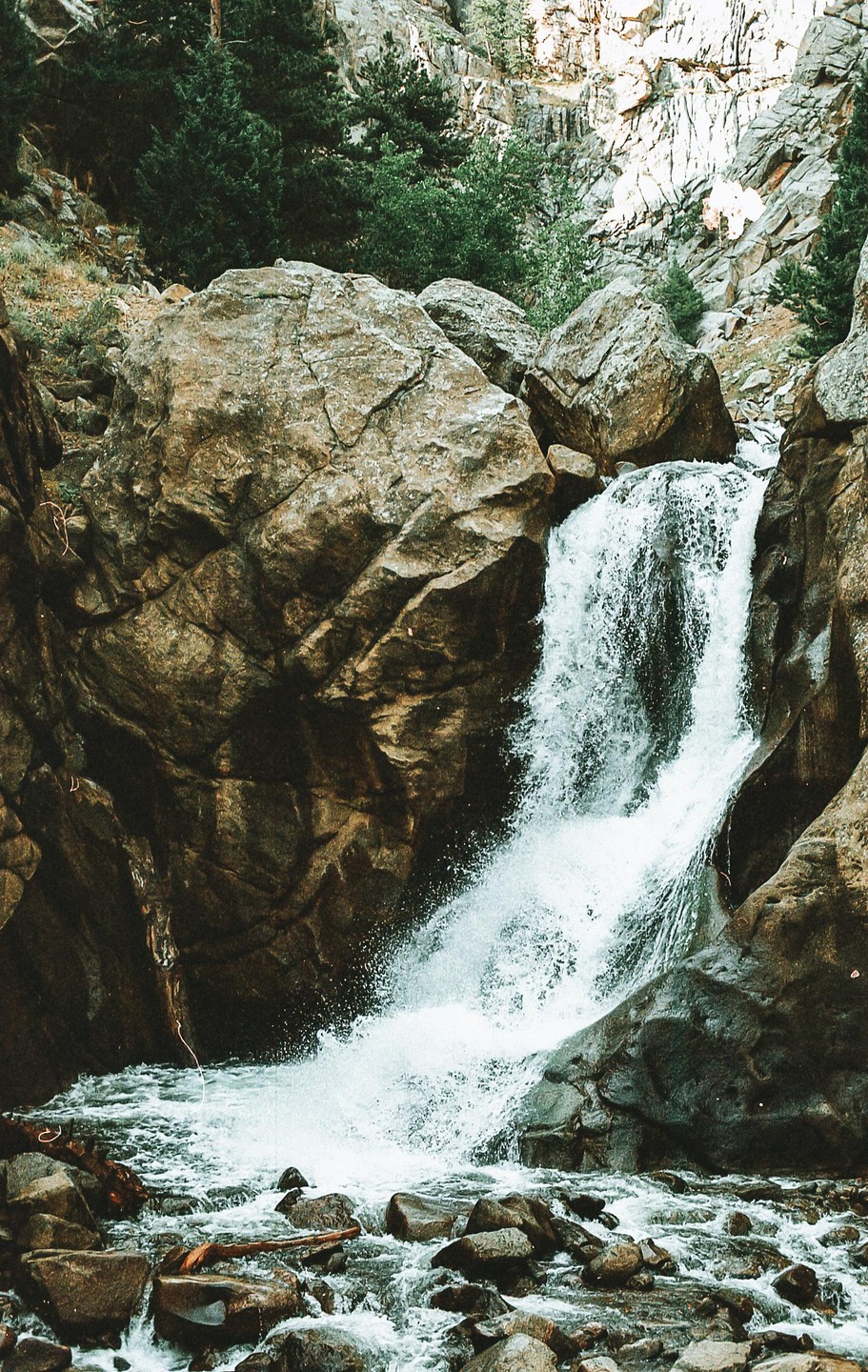 A waterfall cascades over large rocks into a rocky stream surrounded by trees.