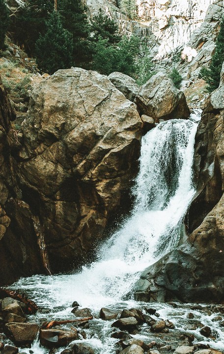 A waterfall cascades over large rocks into a rocky stream surrounded by trees.