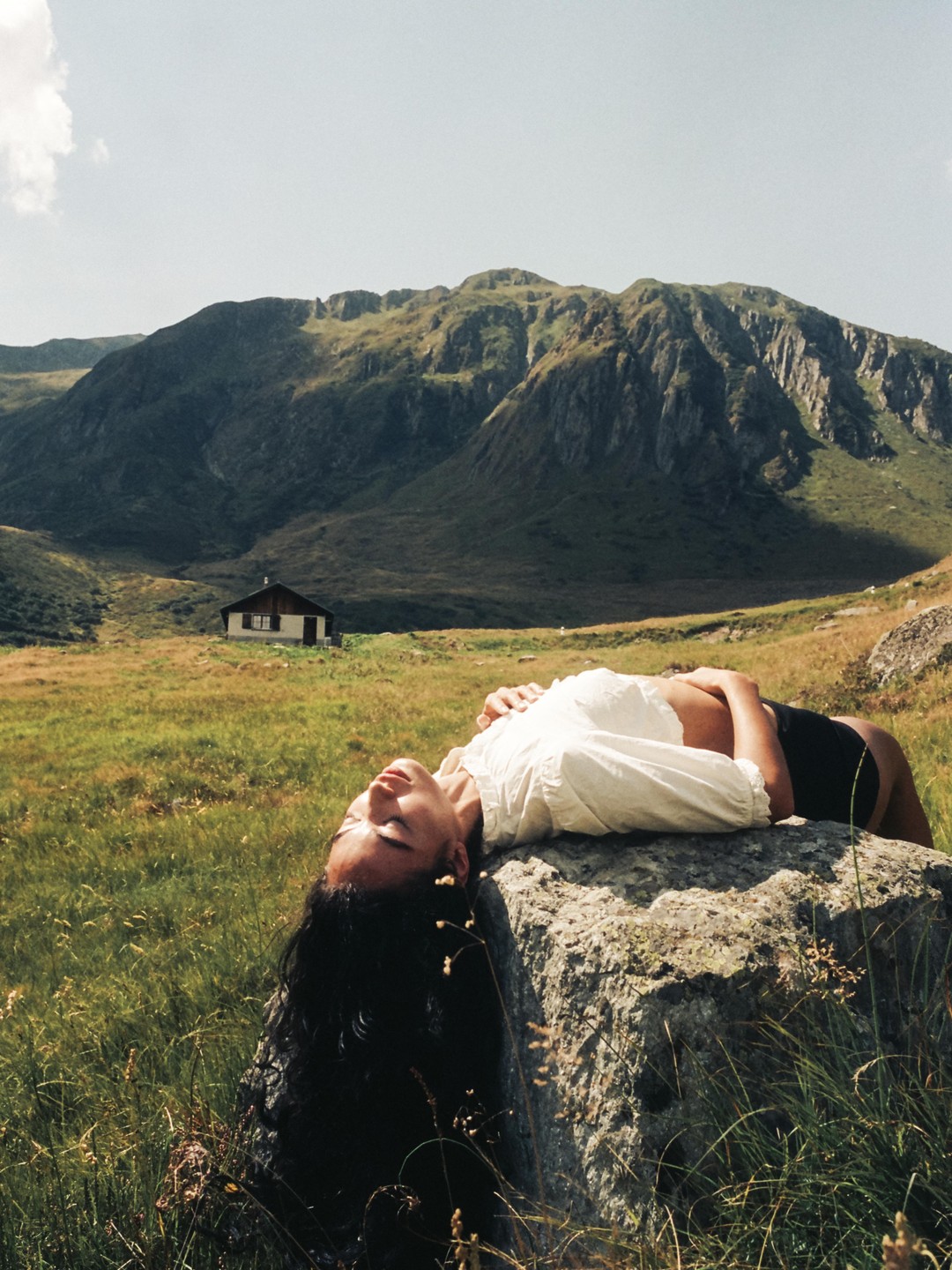 A woman lies on a rock in a grassy field with mountains and a small house in the background.