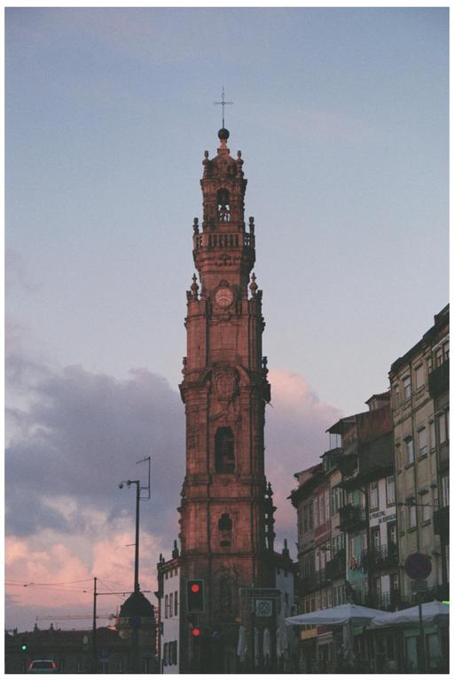 A tall, ornate clock tower stands against a cloudy sky with buildings and streetlights visible on either side.