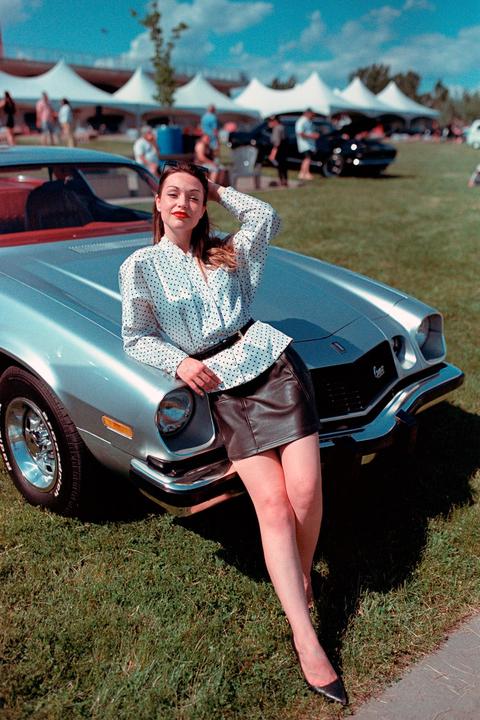 A woman in a polka dot shirt and leather skirt leans on a silver car at an outdoor car show.