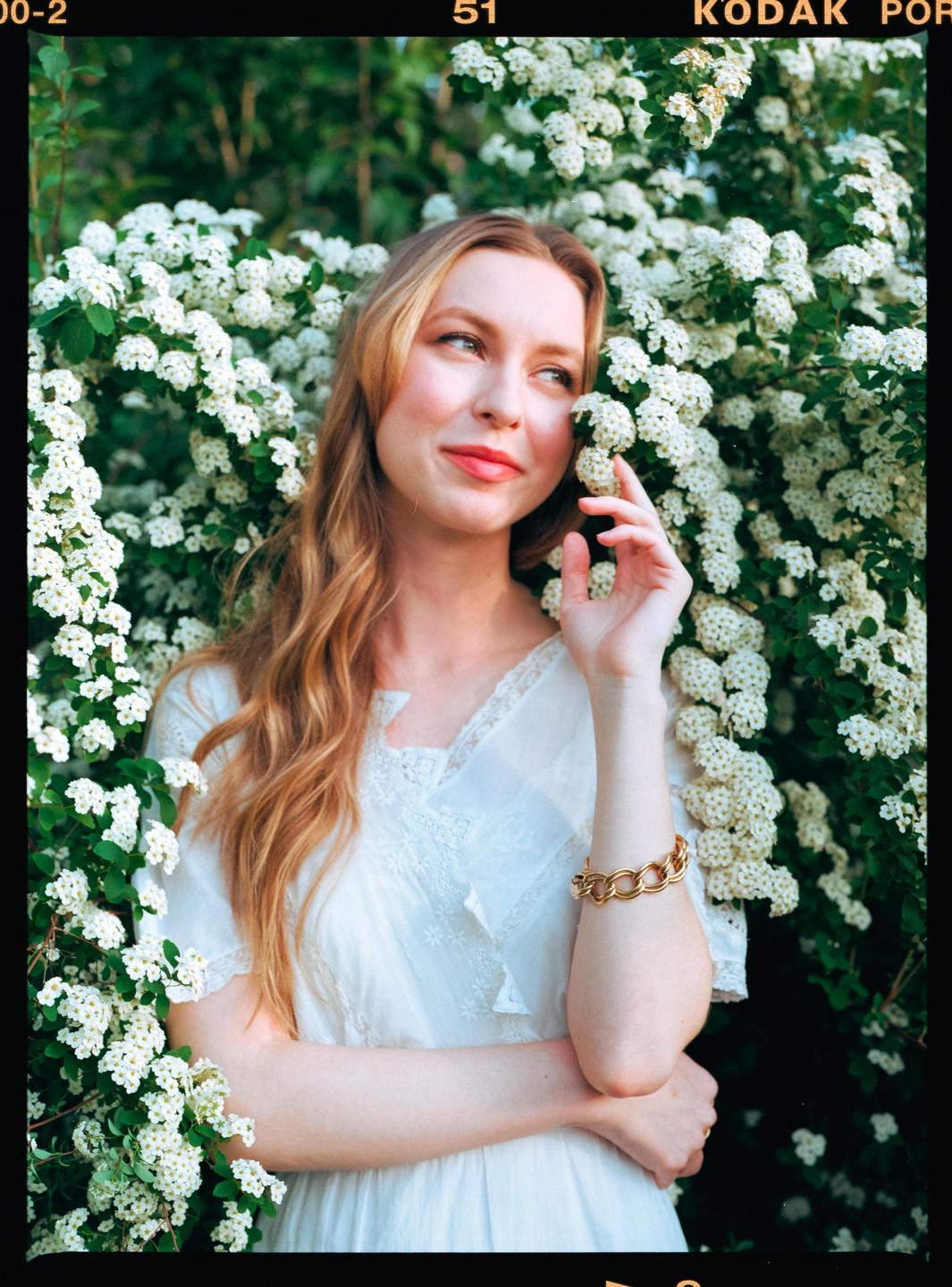 A woman in a white dress stands among white flowering bushes.