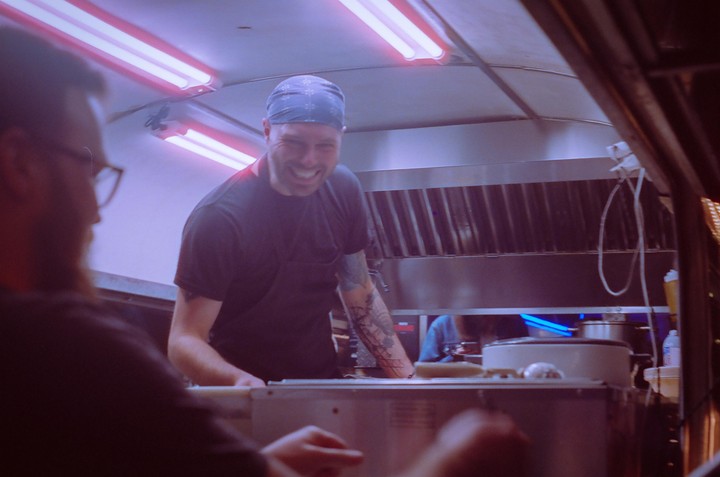 A man with a bandana and a tattooed arm smiles while working in a food truck kitchen.