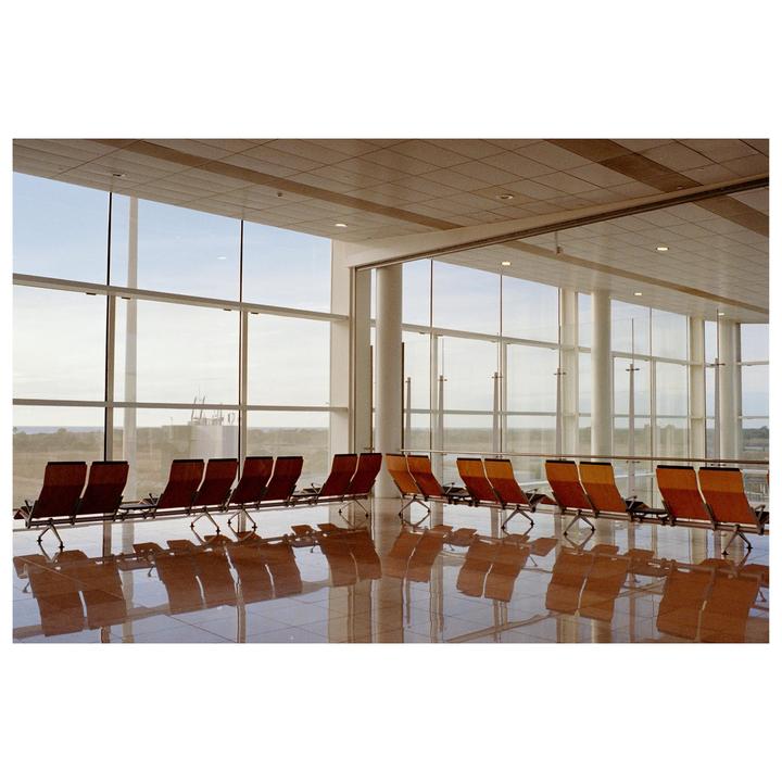 An empty airport waiting area with rows of chairs is illuminated by morning light streaming through large windows.