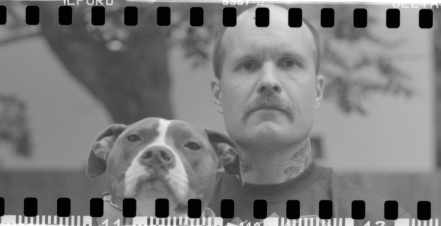 A man with a mustache and neck tattoo poses with a dog.