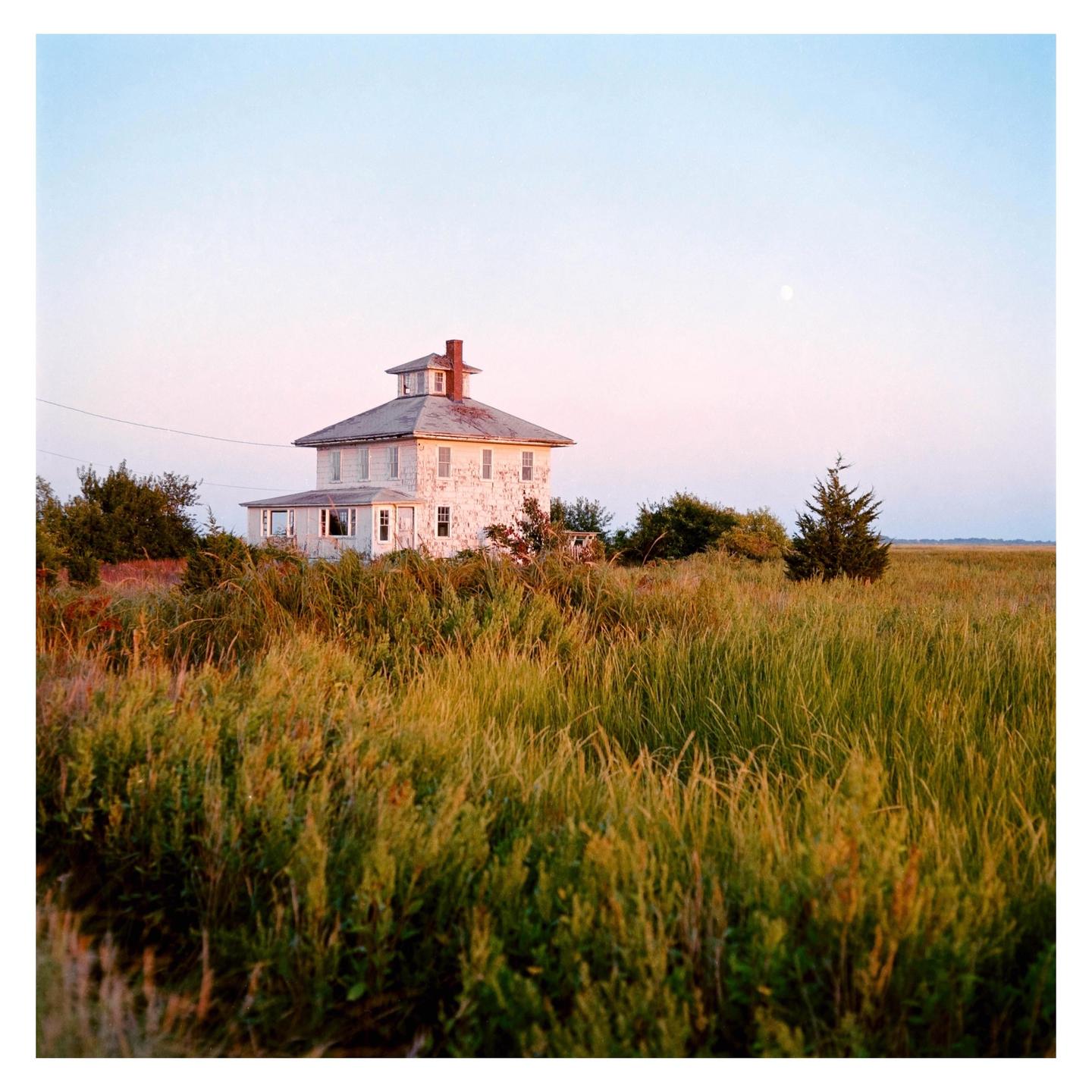 A white house with a red chimney stands in a field of tall grass under a pale sky with a visible moon.