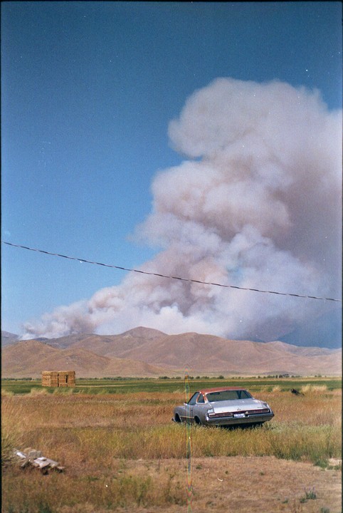 A wildfire burns in the mountains behind a field with a car and hay bales under a blue sky.