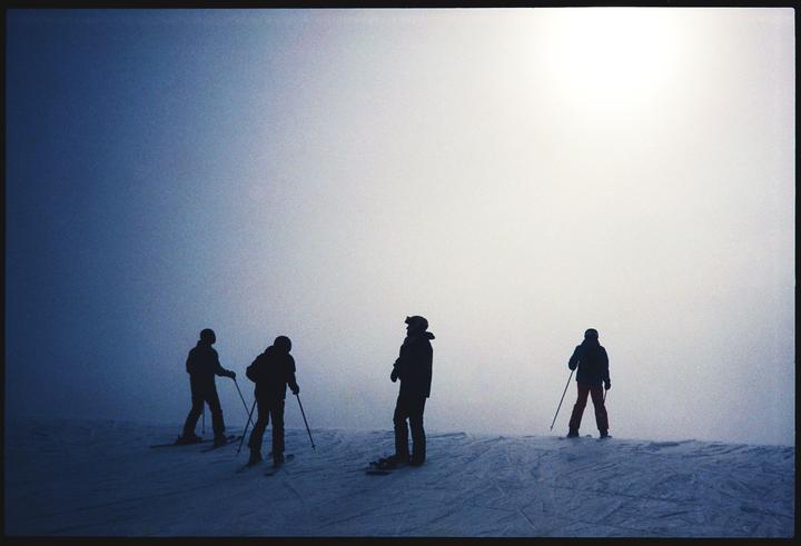 Four skiers are silhouetted against a bright, hazy sky on a snowy slope.