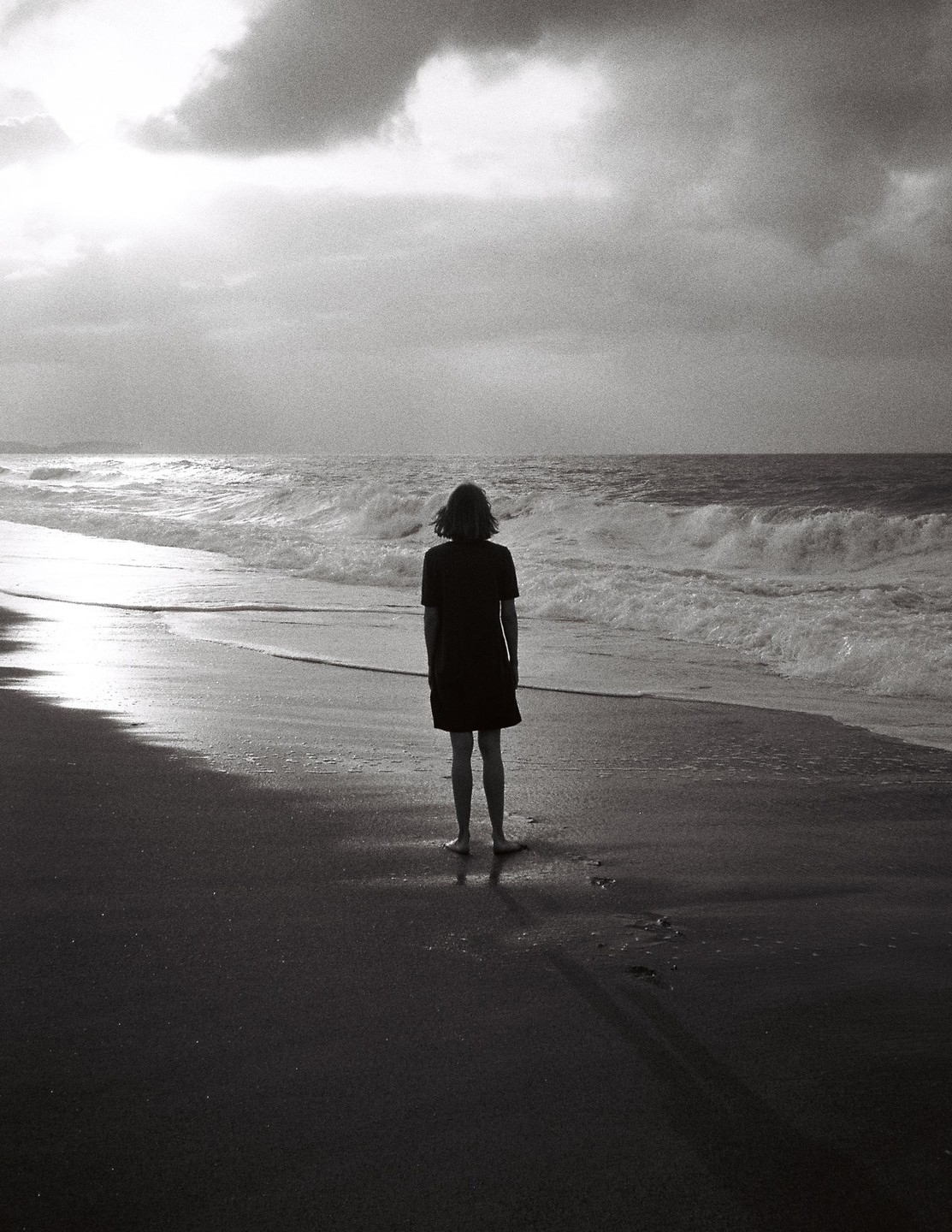 A woman stands on a beach looking out at the ocean under a cloudy sky.