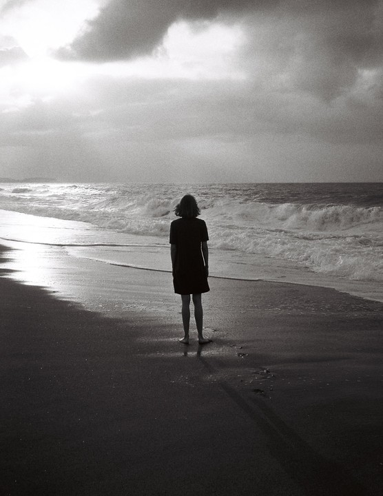 A woman stands on a beach looking out at the ocean under a cloudy sky.