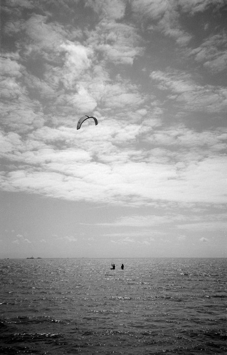 Two people stand in the ocean with a kiteboard kite flying high above them under a cloudy sky.