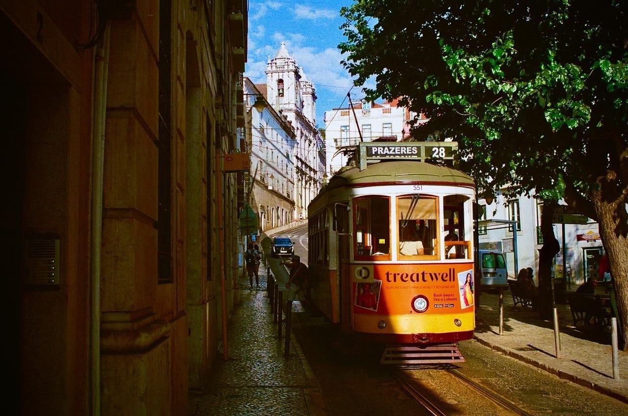A yellow and green tram with "PRAZERES 28" on its destination sign is parked on a narrow, sunlit street in Lisbon, with historic buildings and a large tree framing the scene.