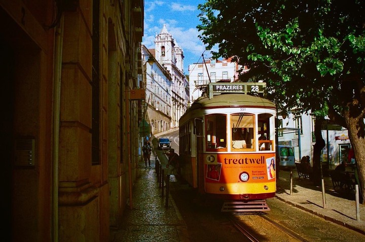 A yellow and green tram with "PRAZERES 28" on its destination sign is parked on a narrow, sunlit street in Lisbon, with historic buildings and a large tree framing the scene.