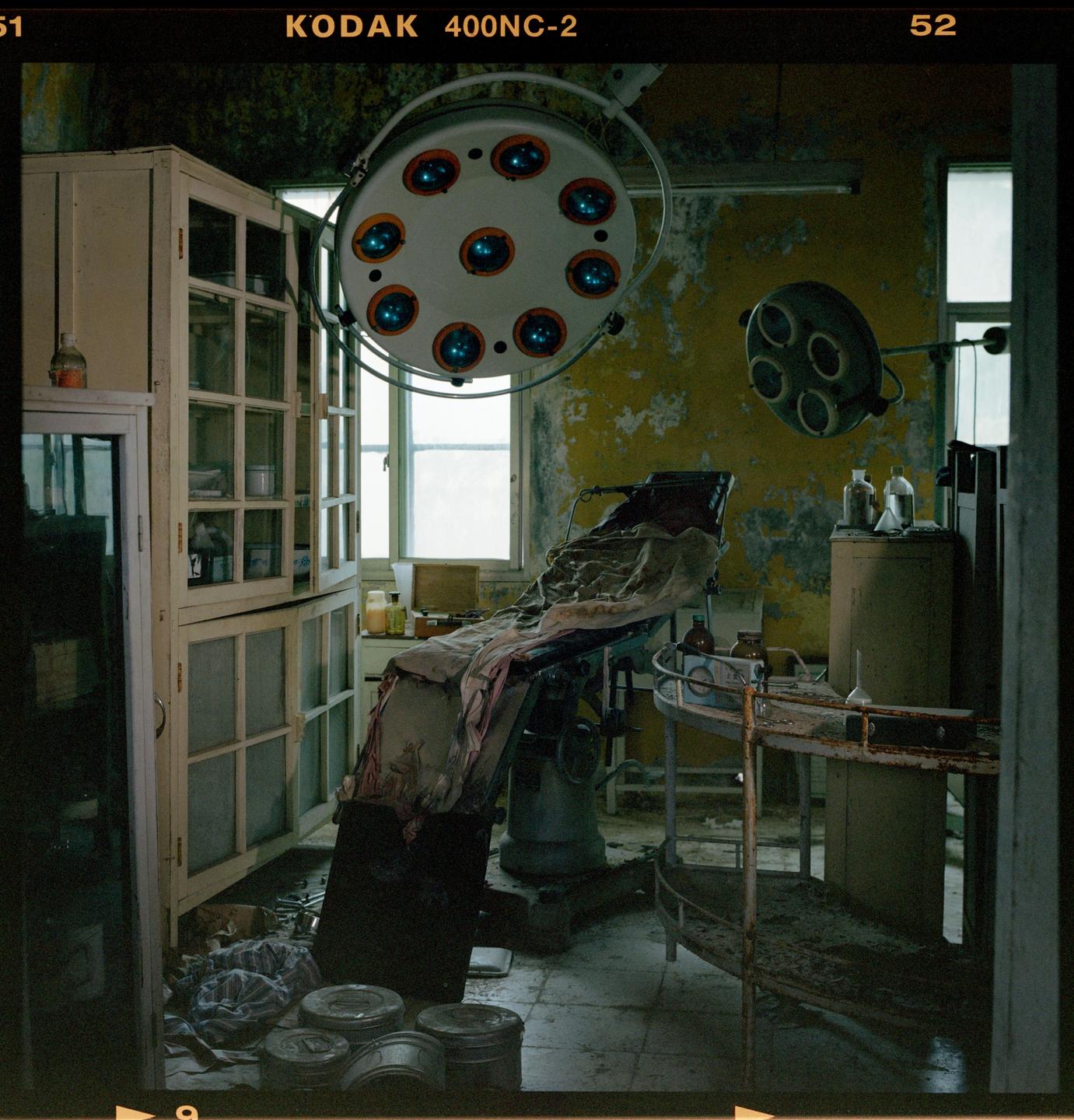An abandoned operating room features a surgical table, medical lights, and cabinets with peeling yellow paint on the walls.