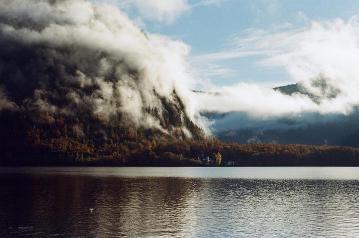 A body of water reflects the sky and a tree-covered mountain partially obscured by clouds.