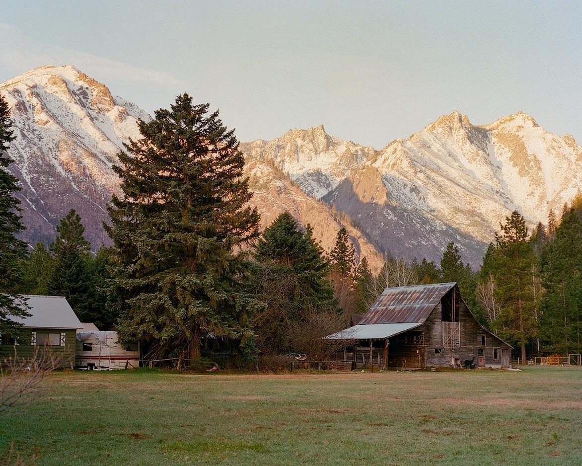 A rustic barn and a house with a camper are nestled in a valley with snow-capped mountains in the background.