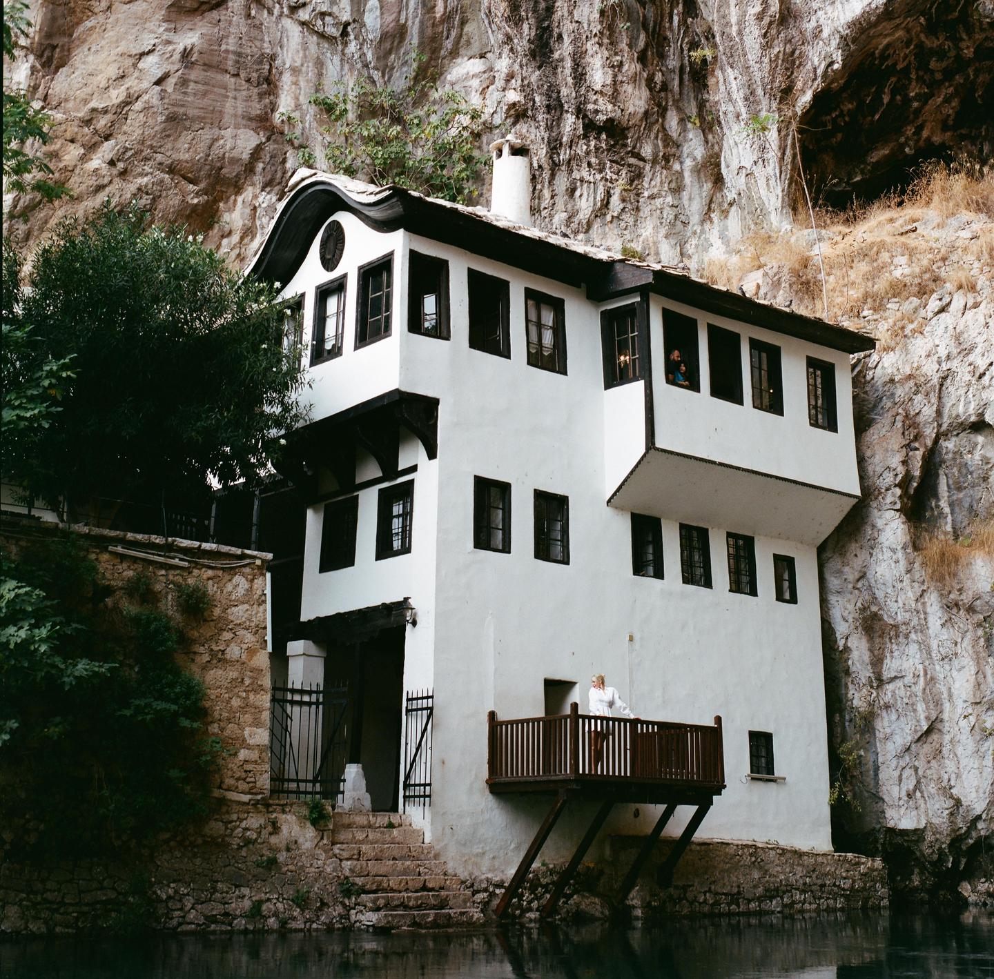 A white house with a dark roof and windows is built into a rocky cliff face above a body of water.