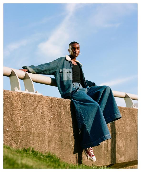 A man wearing a denim jacket and wide-leg jeans sits on a concrete barrier under a blue sky.