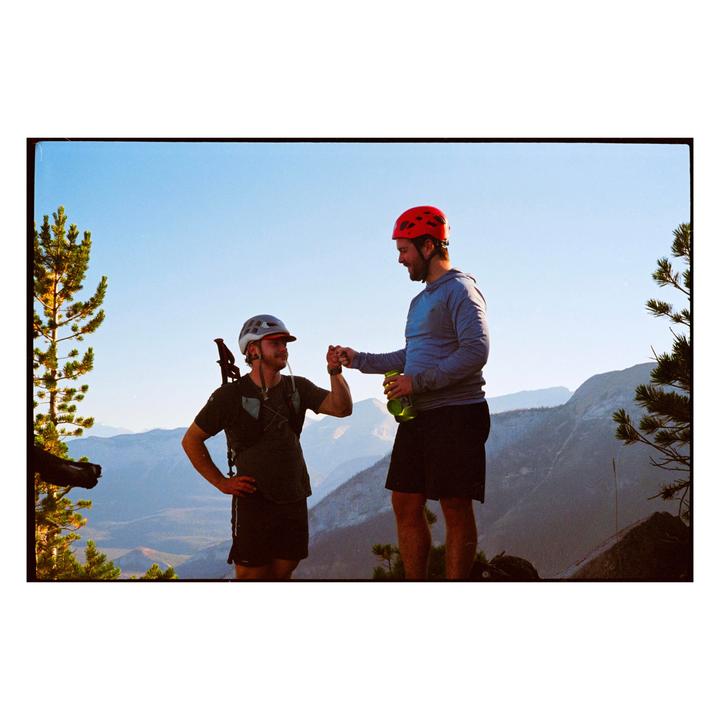Two men in helmets fist bump on a mountain with a vast landscape behind them.