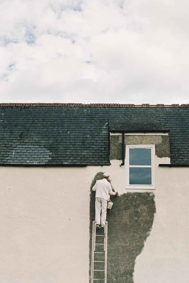 A man on a ladder paints the side of a building with a window and a dark roof.