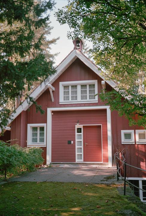 A red wooden house with white trim and a bell on the roof is partially obscured by green foliage.