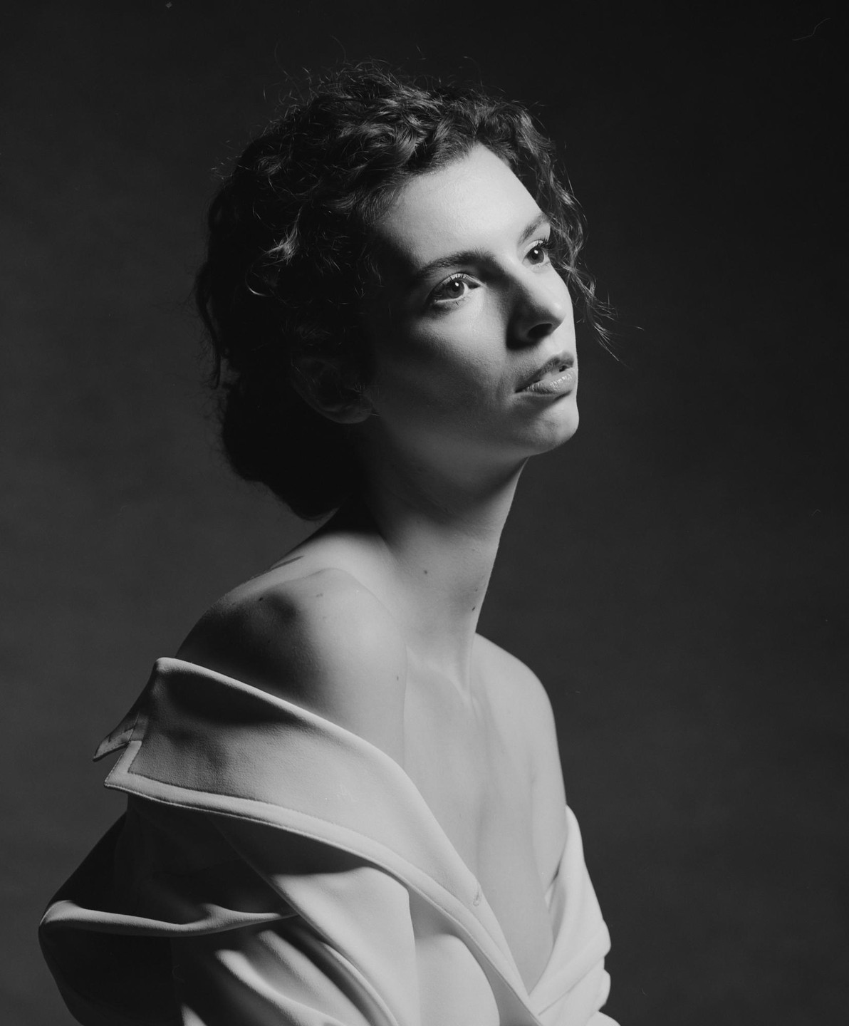 A woman with curly hair and bare shoulders looks up and to the right, wearing a white jacket draped over her arms.