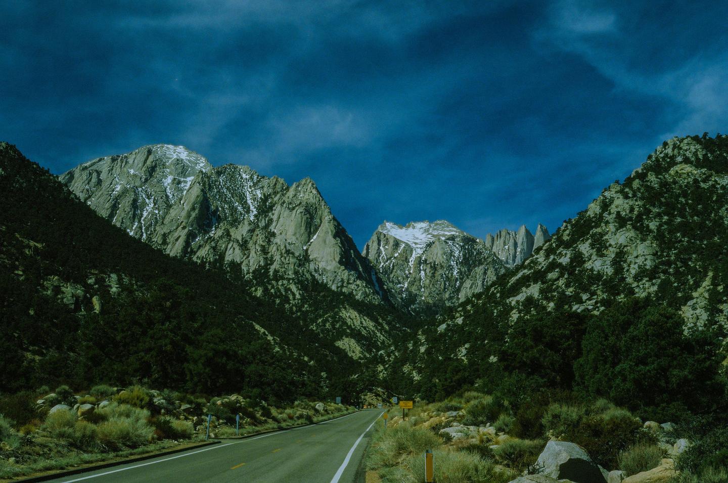 A road leads through a valley with mountains on either side, some with snow-capped peaks.