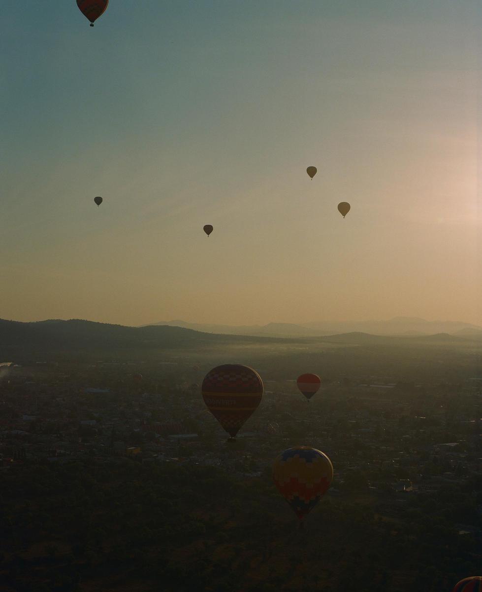 Hot air balloons float above a city at dusk with mountains in the distance.