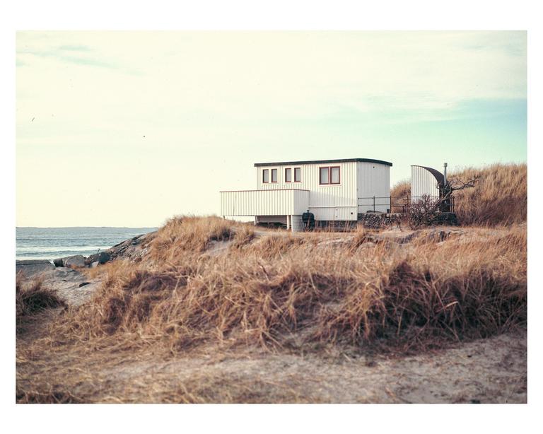 A white house with red window frames sits on a grassy dune overlooking the ocean.
