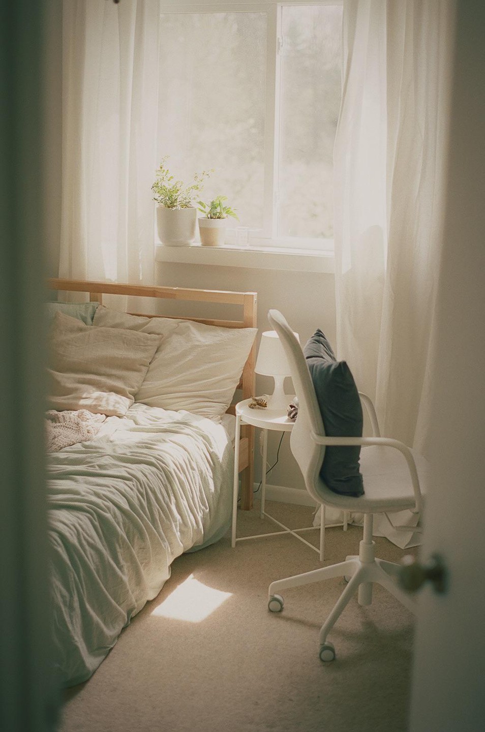 Sunlight streams into a bedroom with a bed, a desk chair, and potted plants on the windowsill.