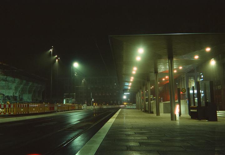 A bus stop with a long covered walkway is illuminated at night next to a wet street with tram tracks.