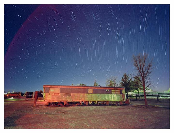 A long exposure captures star trails above a trailer and a bare tree at night.
