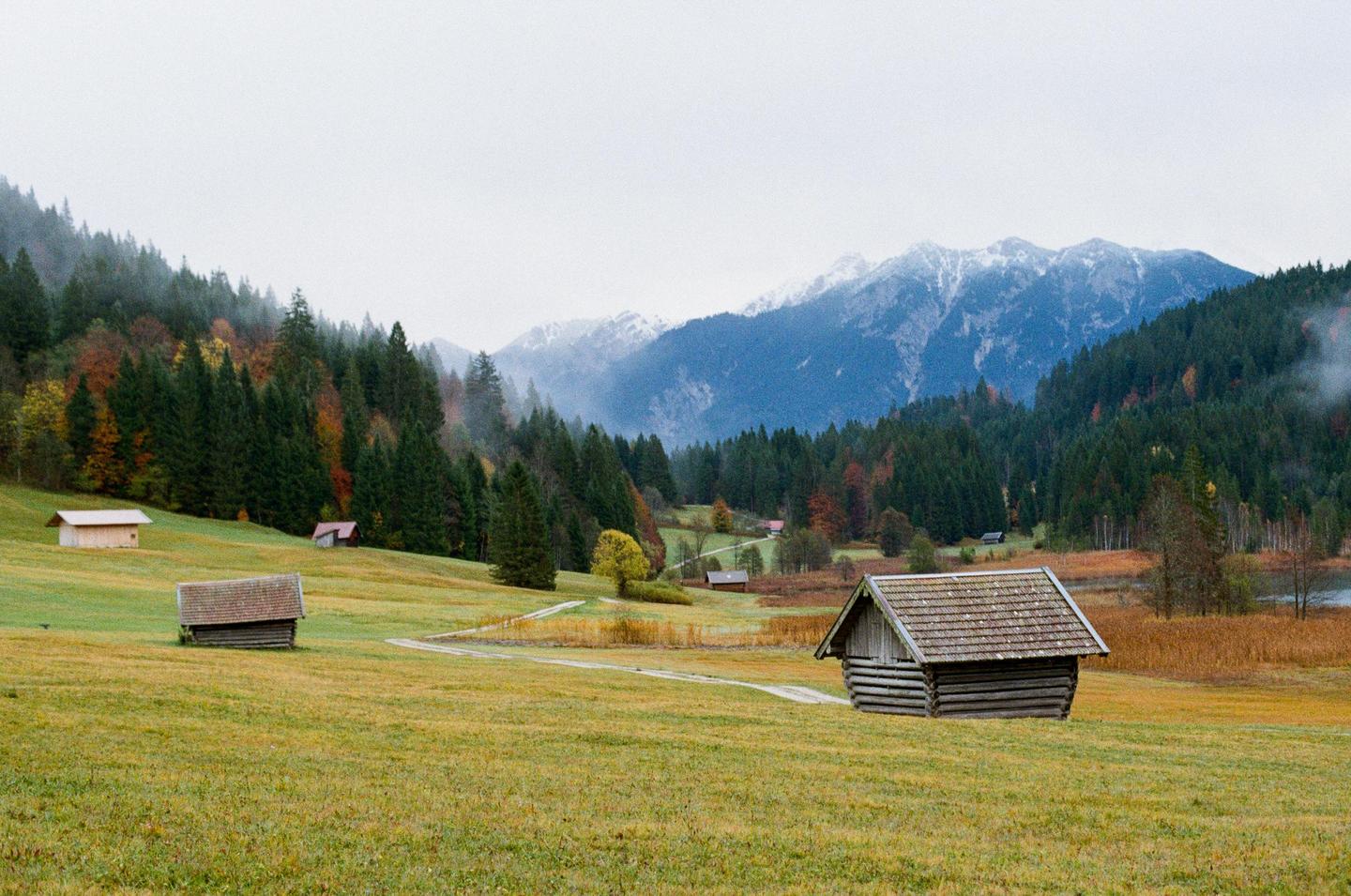 A small wooden cabin sits in a grassy field with mountains and a forest in the background.