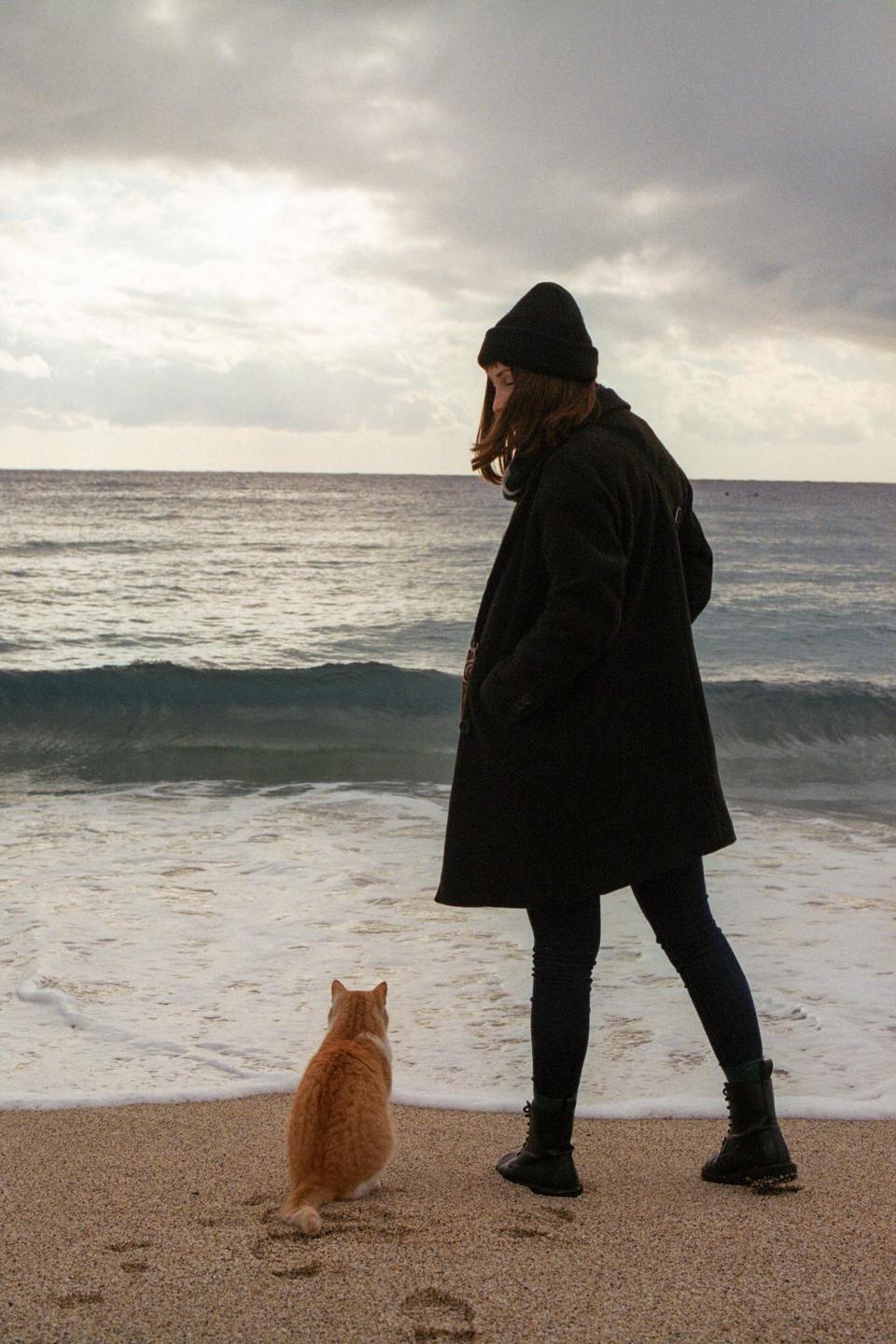 A woman and a cat stand on a sandy beach looking out at the ocean under a cloudy sky.