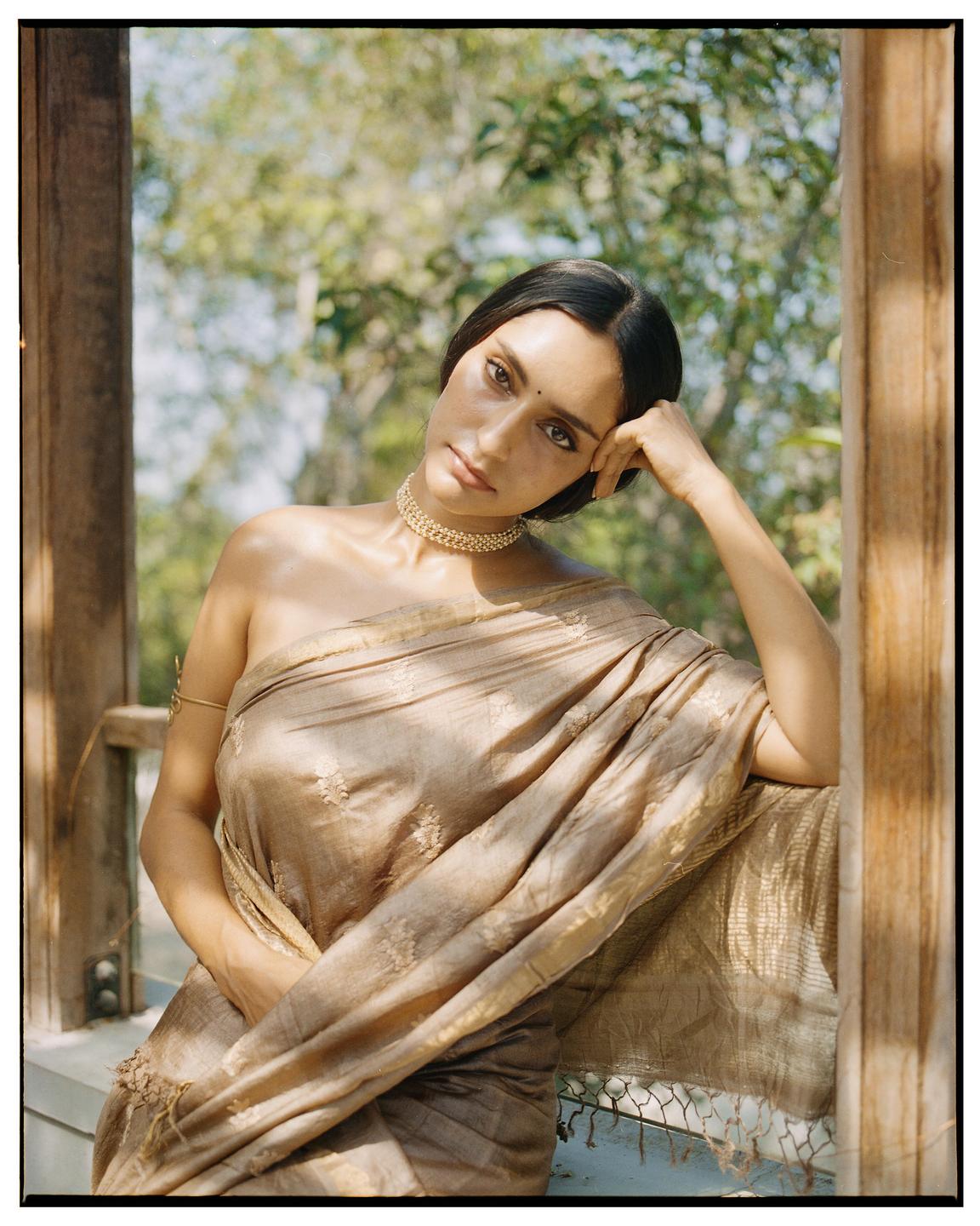 A woman wearing a gold sari and choker necklace poses with her arm resting on a wooden railing.