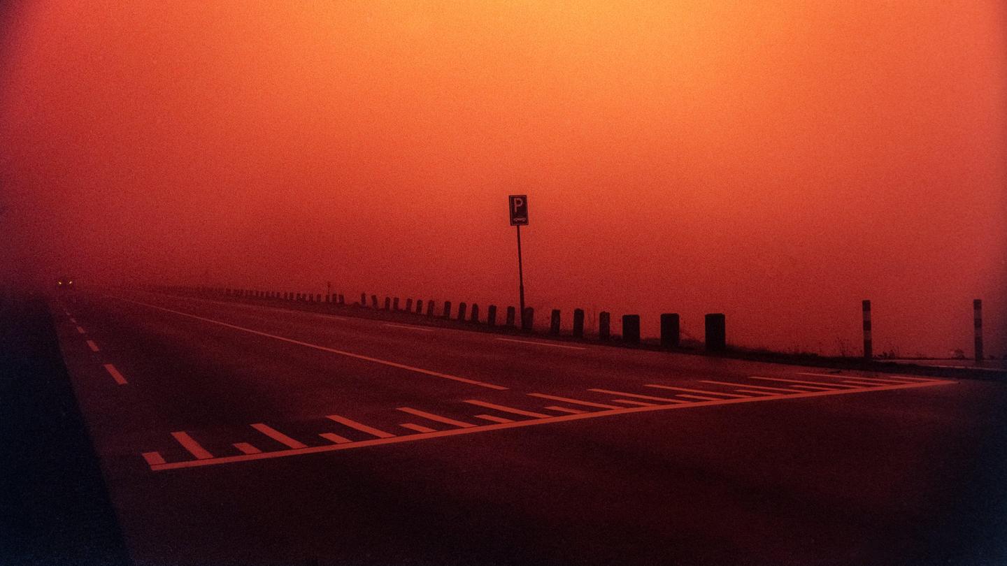 A road with a parking sign and a barrier is shrouded in red fog.