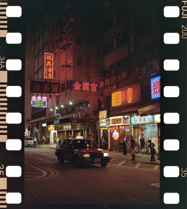 A red taxi drives down a street lined with buildings and neon signs at night in Hong Kong.