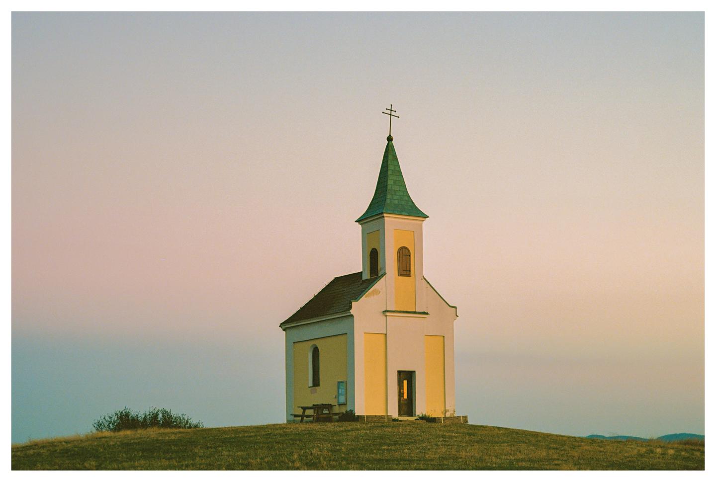 A small chapel with a green roof and a cross on top stands on a grassy hill under a pastel sky.