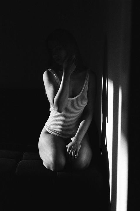 A woman kneels on a couch in a dark room with a beam of light illuminating her and the wall.