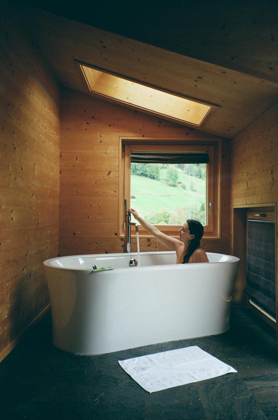 A woman sits in a bathtub filling with water in a wood-paneled bathroom with a view of a green hillside.