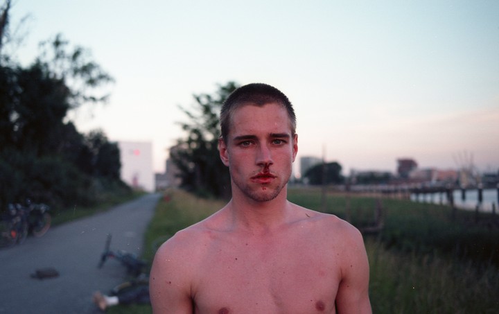 A man with a bloody nose stands shirtless on a path next to a body of water at dusk.