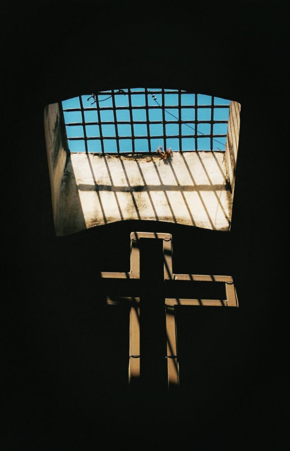 Sunlight streams through a barred window, casting a cross-shaped shadow onto a dark wall.