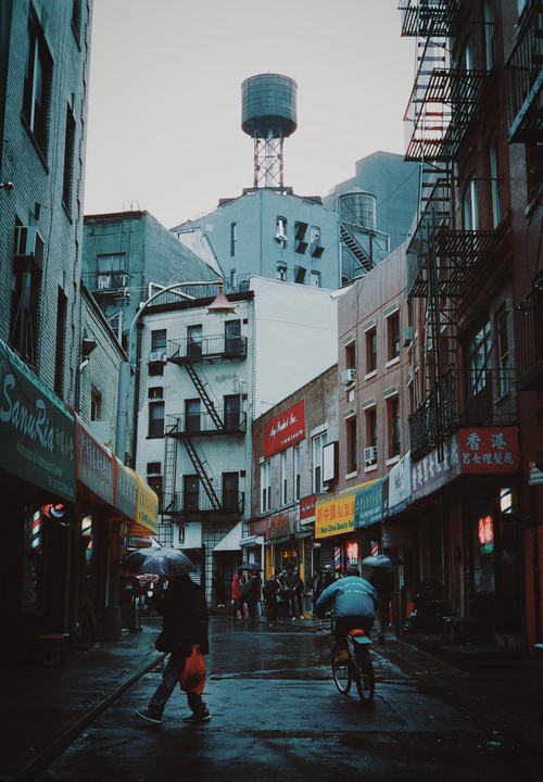 A person with an umbrella walks on a wet street past storefronts and a cyclist, with buildings and water towers in the background.