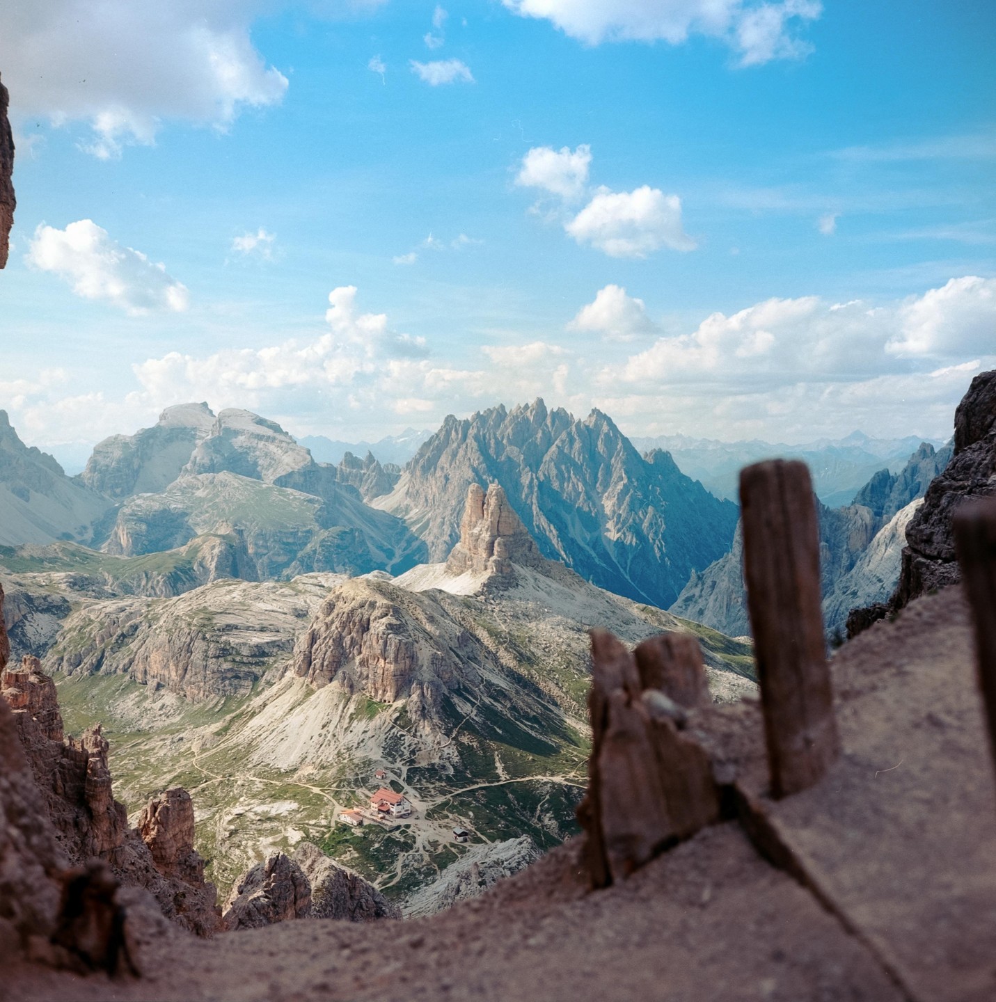 A mountain landscape shows a valley with a building complex and a winding road, surrounded by rocky peaks under a blue sky with clouds.