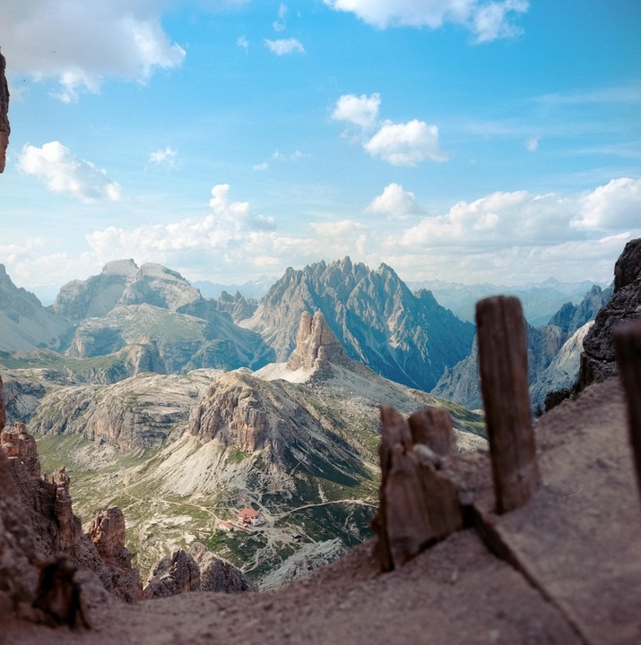 A mountain landscape shows a valley with a building complex and a winding road, surrounded by rocky peaks under a blue sky with clouds.